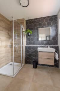 Ein modernes Badezimmer mit einer Duschnische mit Glaswand und Duschtasse und einem Fliesenmix aus Holzoptikfliesen im Duschbereich und schwarzen Wabenfliesen an der Wand mit Waschtisch von HARTL HAUS.
