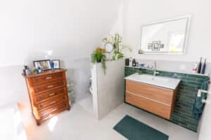 Ein Badezimmer mit Dachschräge, großformatigen hellgrauen Fliesen in Steinoptik, glänzenden grünen Stabfliesen, einem modernen Waschtisch und einem WC mit raumtrennender Zwischenwand, Badewanne und antiker Kommode von HARTL HAUS.