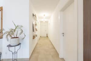 Ein heller Flur im Obergeschoss eines Hauses mit beigen keramischen Bodenfliesen, dekorativen Bilderleisten und harmonierenden Pflanzgefäßen und Accessoires von HARTL HAUS.