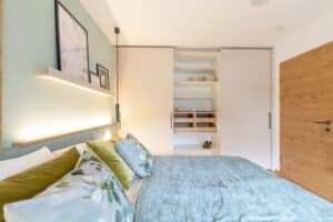 Ein Schlaf- bzw. Gästezimmer mit einem Doppelbett mit gepolstereter Rückenlehne in meerblauem Samt, passender Bettwäsche und einem weissen Einbaukleiderschrank der viel Stauraum bietet und für eine kompakte Raumgestaltung sorgt - von HARTL HAUS.