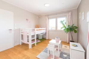 Ein liebevoll gestaltetes Kinderzimmer mit Hochbett, rosa Akzenten, Spielzeug, Kindertisch mit zwei Stühlen, Zimmerpflanze und beigen Wänden von HARTL HAUS.