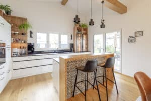 Eine moderne Einbauküche mit Kochinsel mit integrierter Bar, stilvollen Barhockern und Blick auf die Terrasse durch Hebeschiebetüren von HARTL HAUS.