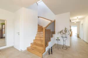 Ein heller Vorraum mit eleganter Holztreppe, Stabgeländer mit geschwungenem Holzhandlauf, großformatigen Bodenfliesen und stilvoller Raumgestaltung von HARTL HAUS.