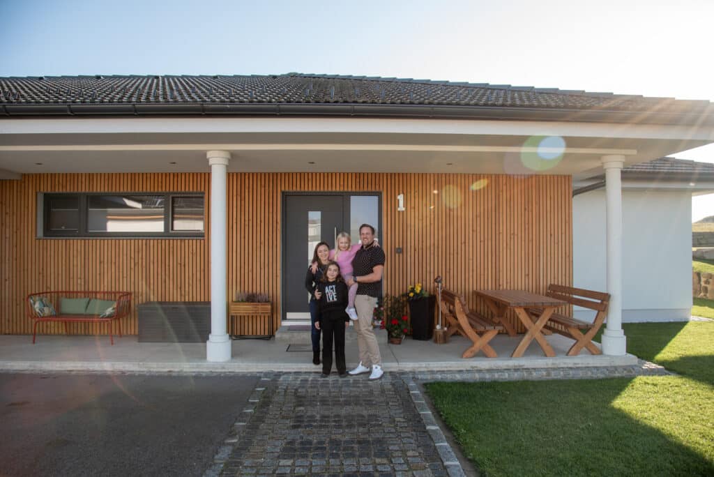 Eine junge Familie vor einem modernen Bungalow von HARTL HAUS mit Veranda bei sonnigem Wetter.