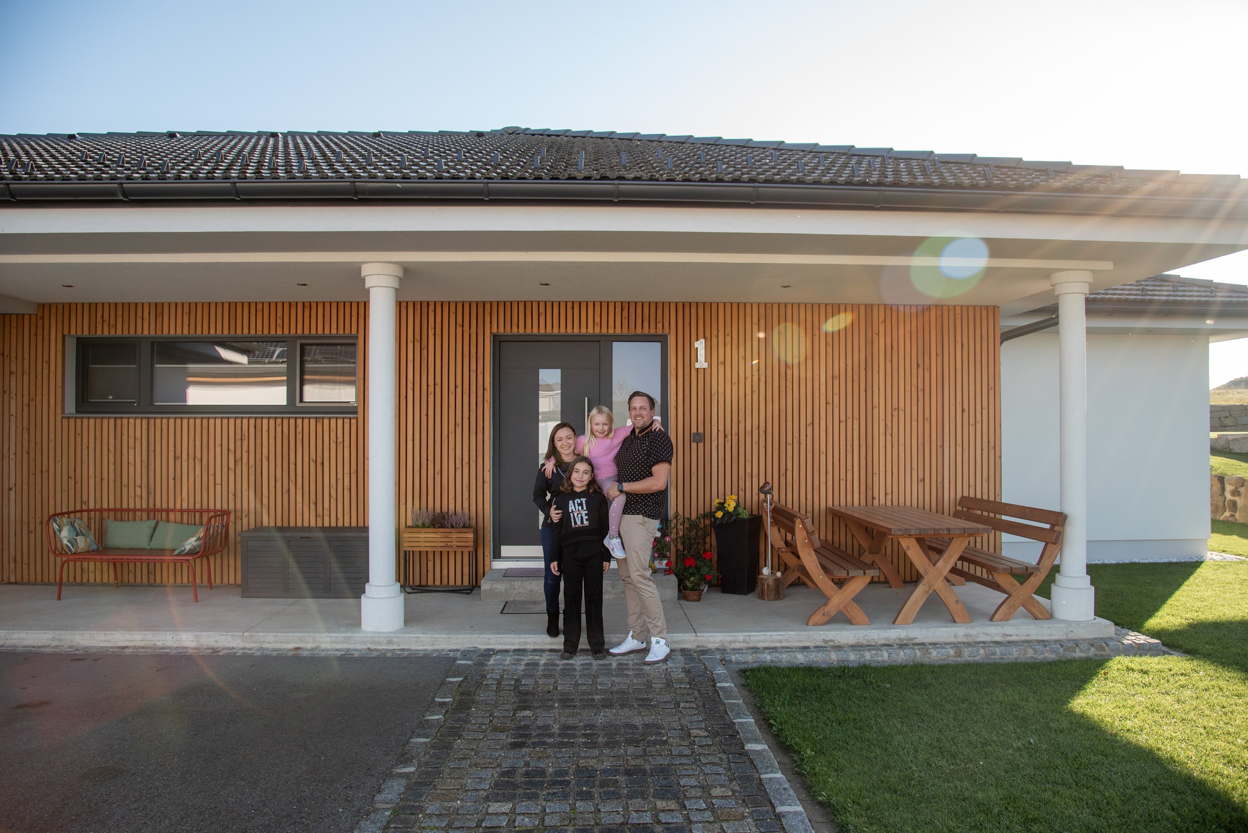 Eine junge Familie vor einem modernen Bungalow von HARTL HAUS mit Veranda bei sonnigem Wetter.