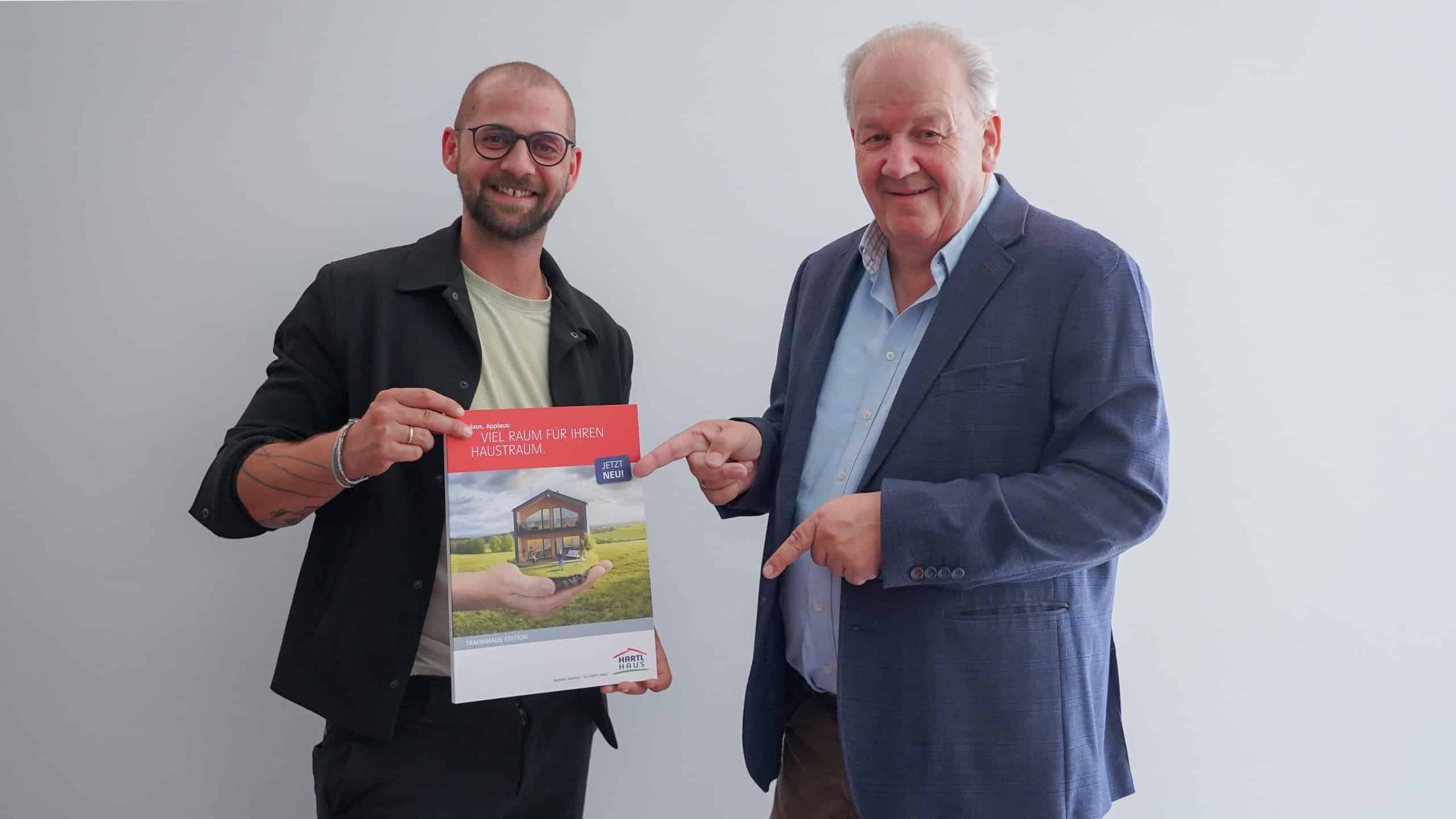Herr Drexl und Herr Müller posieren mit dem Traumhauskatalog von HARTL HAUS der über 40 Hausinspirationen enthält.