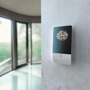 Eine Jablotron JA-116E Natus Touchscreen-Bedieneinheit ist als Wandmodul auf einer Sichtbetonwand montiert dargestellt und zeigt einen aktivierten Bildschirmschoner zur modernen Steuerung des Alarmsystems von Heimdall Alarmsysteme.