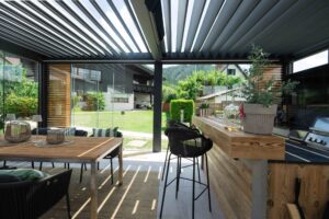 Innenbereich einer Pergola mit Lamaxa L60 Lamellendach mit Bar, Outdoorküche, Glasschiebetüren und äußeren Holzschiebeelementen die zur Beschattung - ein perfekter Sommer- bzw. Wintergarten von Warema, gezeigt von HELLSAN.