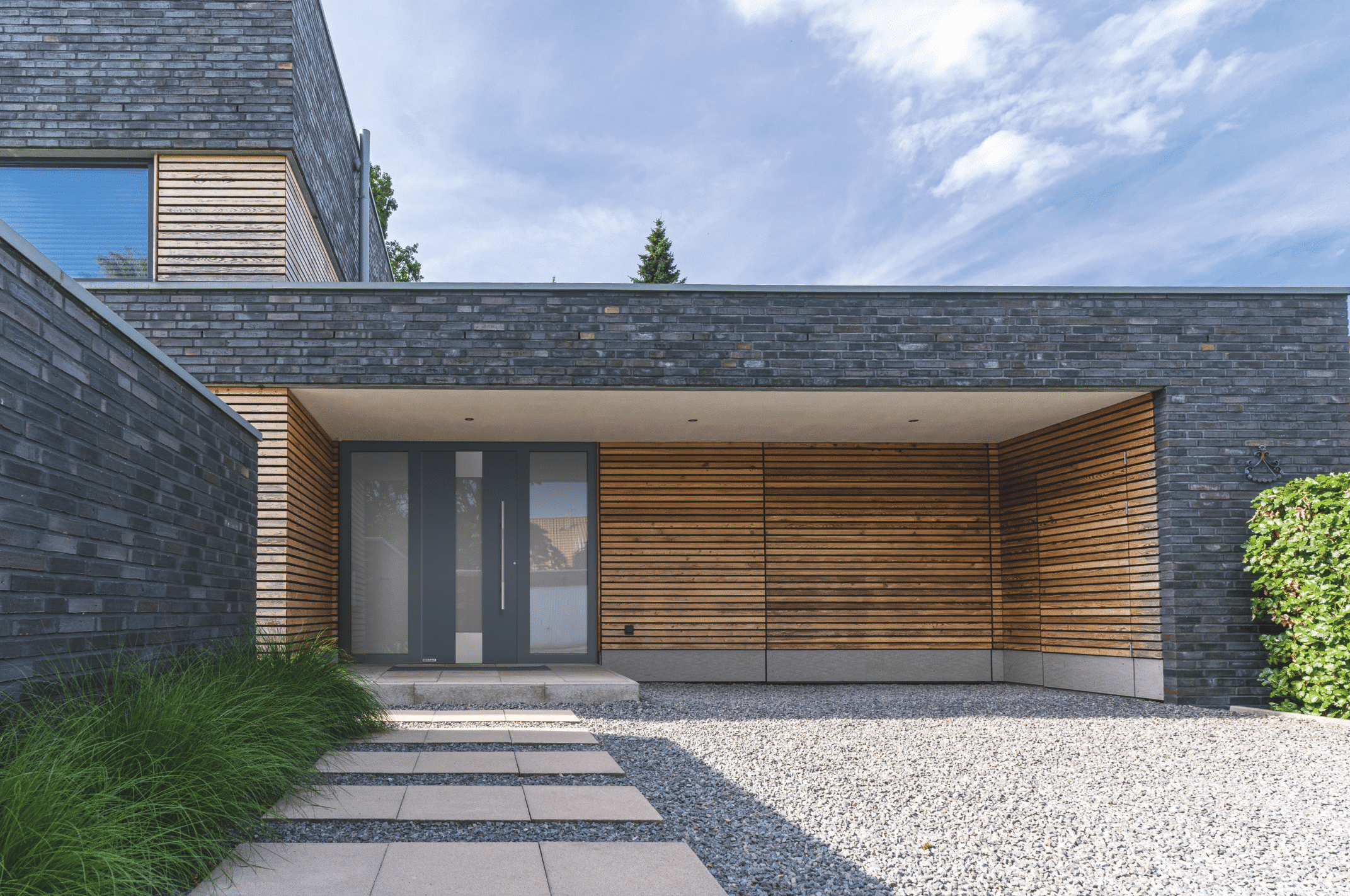 Hörmann Austria zeigt ein modernes Haus mit Holzverkleidung, Begrünung im Vorgarten und Steinplatten als Zugang zur grauen Haustüre mit silbernem Griff und Sichtfenstern.