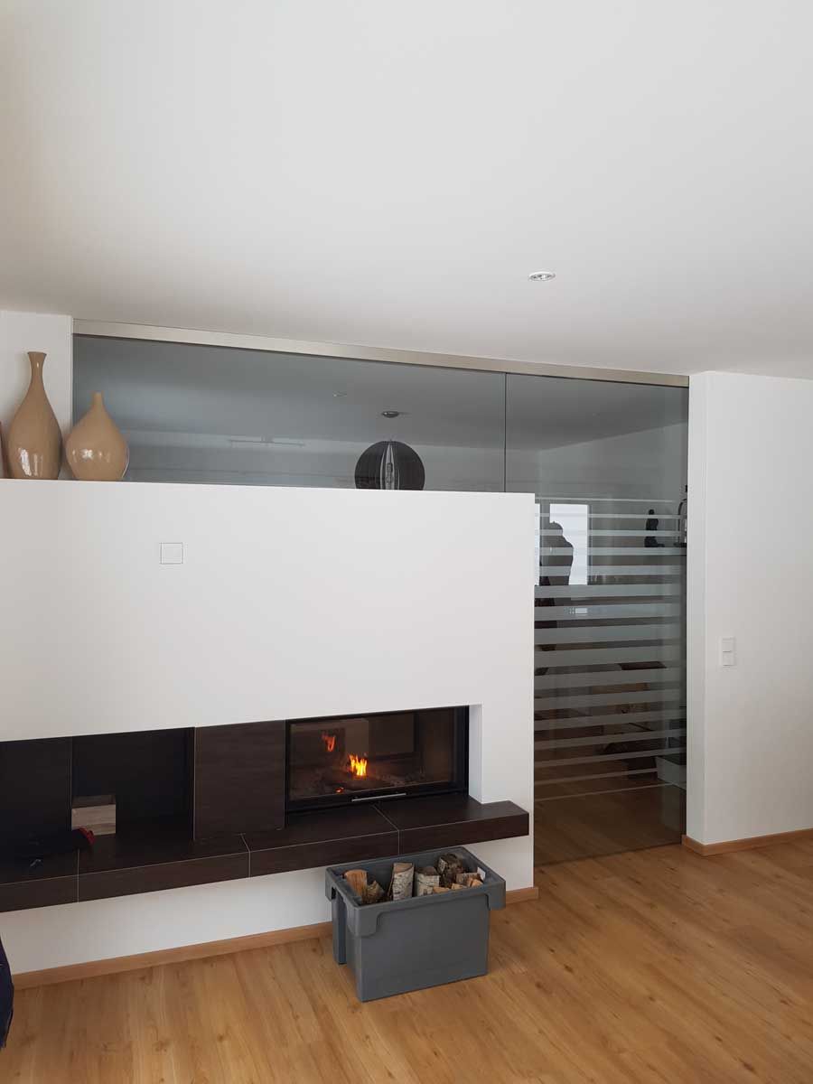 Wohnraum mit modernem, gemauerten, raumtrennenden Kamin dahinter eine Raumtrennung aus Glas mit Durchgangstüre und satiniertem Glasmuster von Glaserei Hohl.