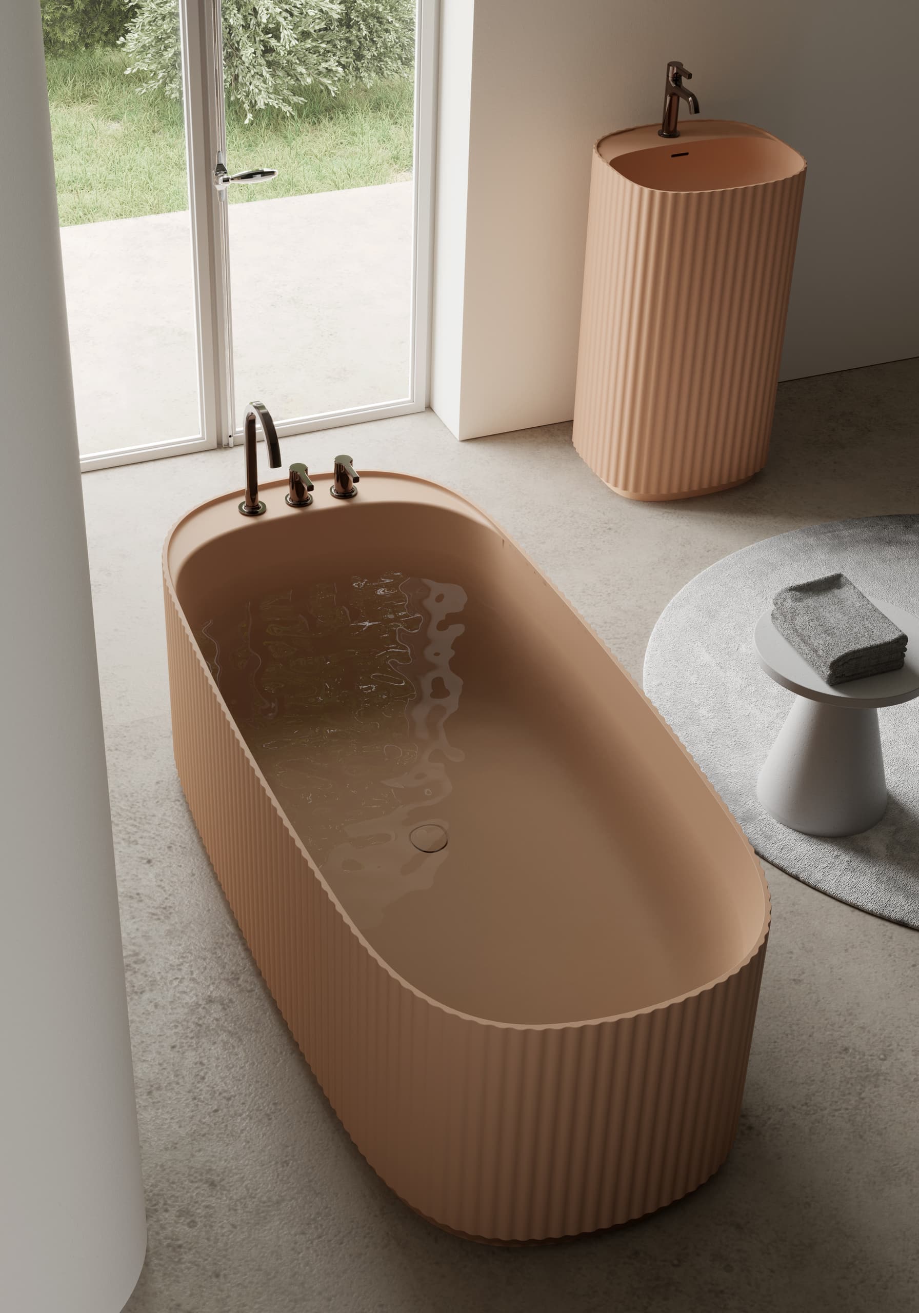 Freistehende ovale Badewanne und Waschsäule mit geriffelter Oberfläche in mattem Lachsrosa, integrierten Armaturen in Bronzeoptik, auf Betonboden mit grauem Beistelltisch und passendem Rundteppich von Disenia.
