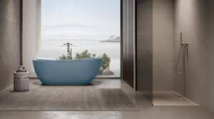 Freistehende ovale Badewanne in Hellblau mit zentral positionierter Armatur, vor raumhoher Fensterfront mit Seeblick, auf Betonboden mit Teppich, Beistelltisch und einer zusätzlichen Walk-in-Duschnische von Disenia.