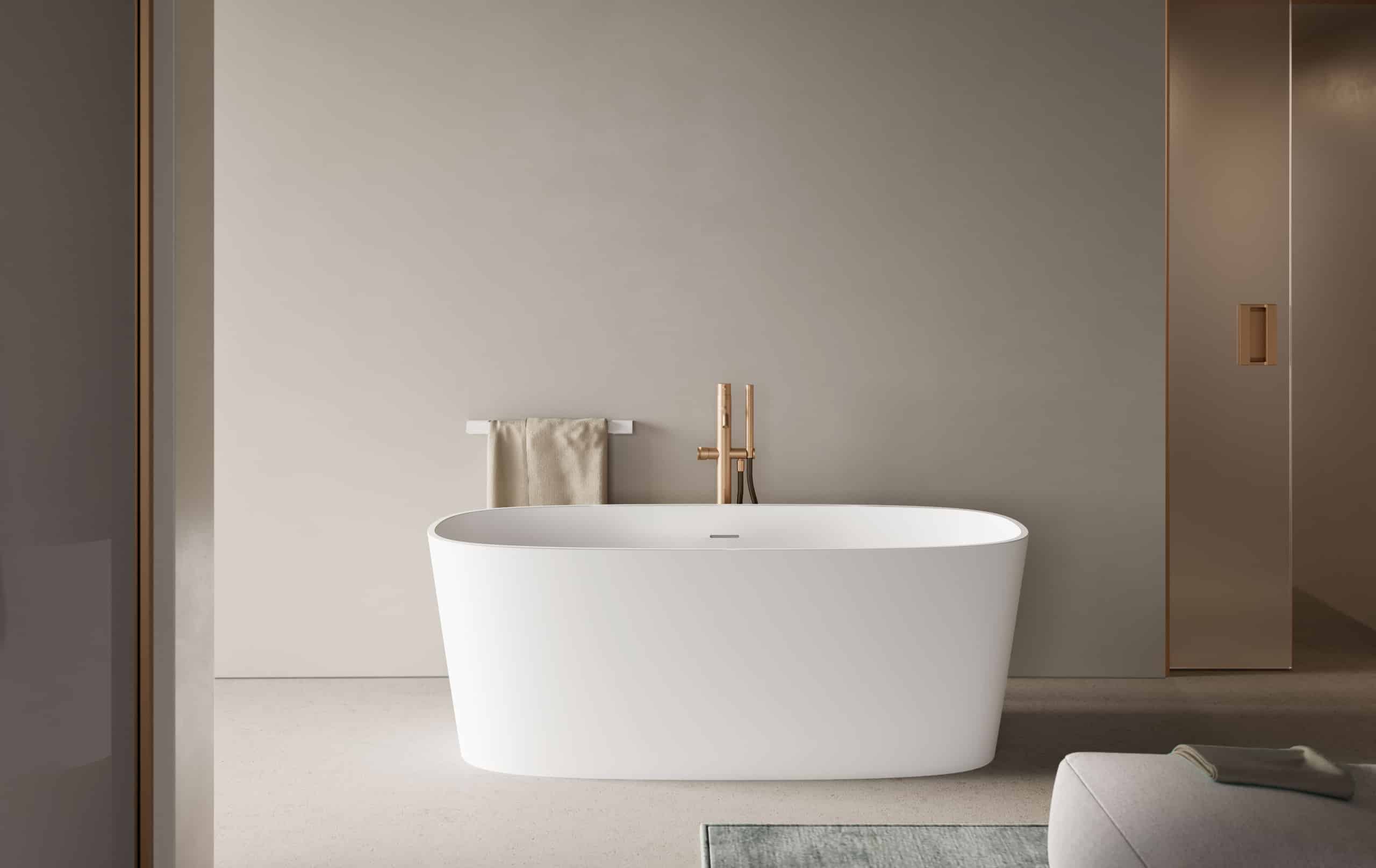 Freistehende ovale Badewanne in strukturiertem, matten Weiß, ergänzt durch eine freistehende Armatur in Roségold mit Handbrause und einem Handtuchhalter vor beiger Wand von Disenia.