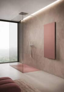 Disenia von Idea Group präsentiert eine an zwei Seiten offene Dusche mit einer Glaswand abgetrennt und mit Armatur und Handdusche aus chrom. Daneben ist ein Heizpaneel in altrosa an der Wand monitert.