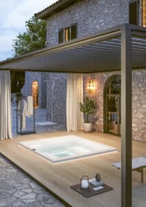 Eine Terrasse mit Pergola, seitlichen Vorhängen und Lamellendach und einer Unterbau-Version des Whirlpools "Helba 240" in Weiss mit Nackenstützen bzw. Einstiegshilfen oder Sitzflächen am Rand mit matter Oberfläche von Disenia.