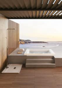 Der rechteckige Whirlpool "Helba 210" in der Unterbau-Version auf einer riesigen Terrasse mit Lamellendach, Outdoordusche und Glasgeländer - hineingebaut in ein Podest in Steinoptik mit Treppen und Sitzbereich, von Disenia.