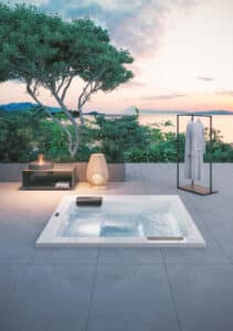 Terrasse mit XL-Steinfliesen und einer Unterbauversion des Outdoor-Whirlpools "Kyra" in Weiss mit schwarzen Nackenstützen bzw. Einstiegshilfen oder Sitzflächen am Rand mit matter Oberfläche und Touchscreen-Panel umgeben von einem stummen Diener, einer Stehlampe und einem Beistelltisch mit Feuerstelle von Disenia.