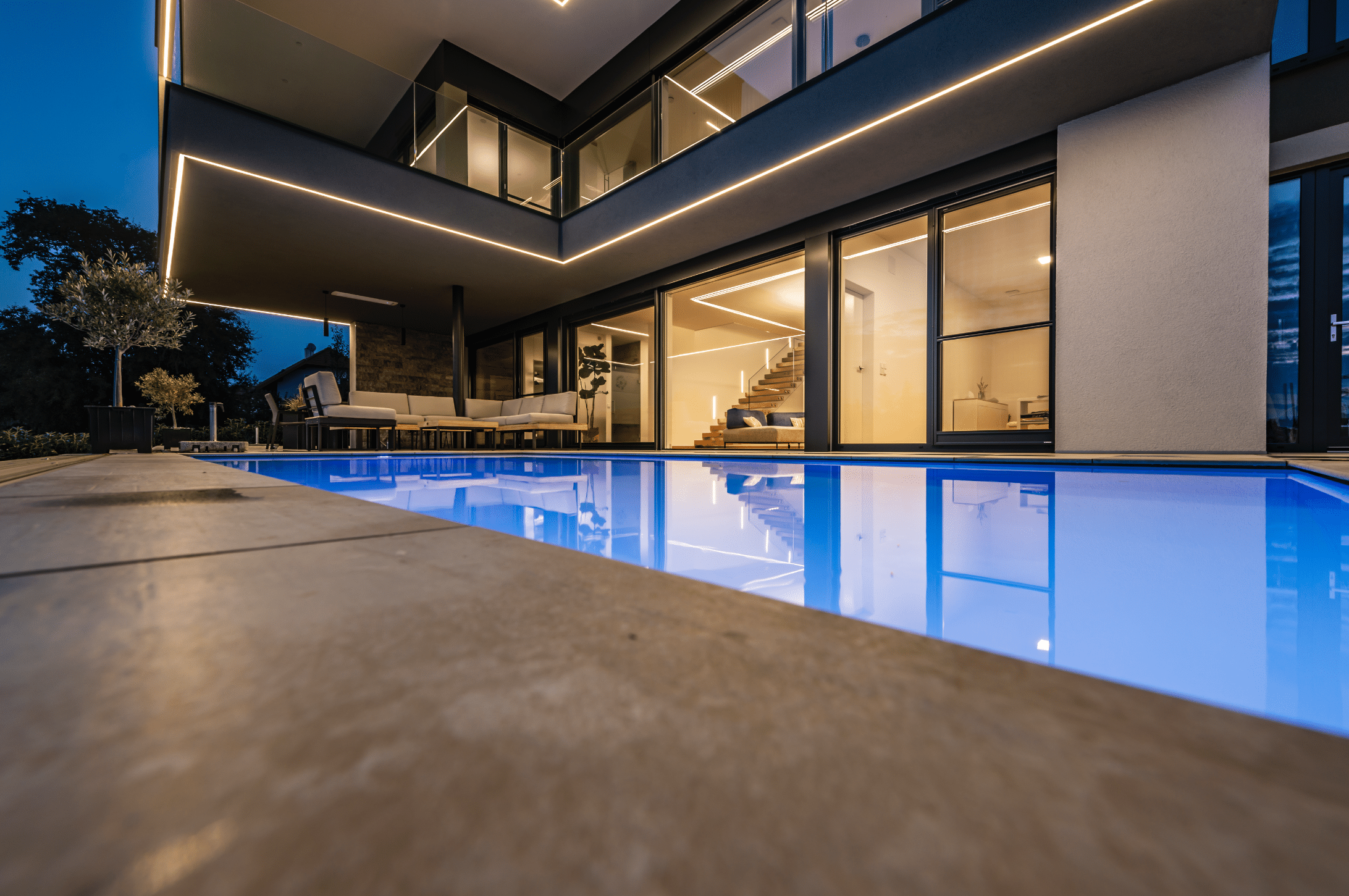 Modernes Flachdach-Wohnhaus mit Glasgeländer Balkon, großer Terrasse mit Pool und vielen raumhohen Fenstern und Glasschiebetüren von Internorm bei Nacht.