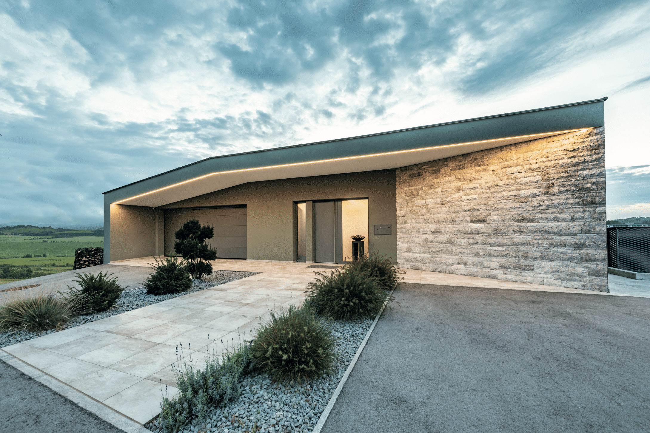 Internorm zeigt modernen Bungalow mit asymetrischem Dach und hellgrauem Garagentor und Außentüre mit Milchglas-Seitenelementen und senkrechter Metallgriffleiste.