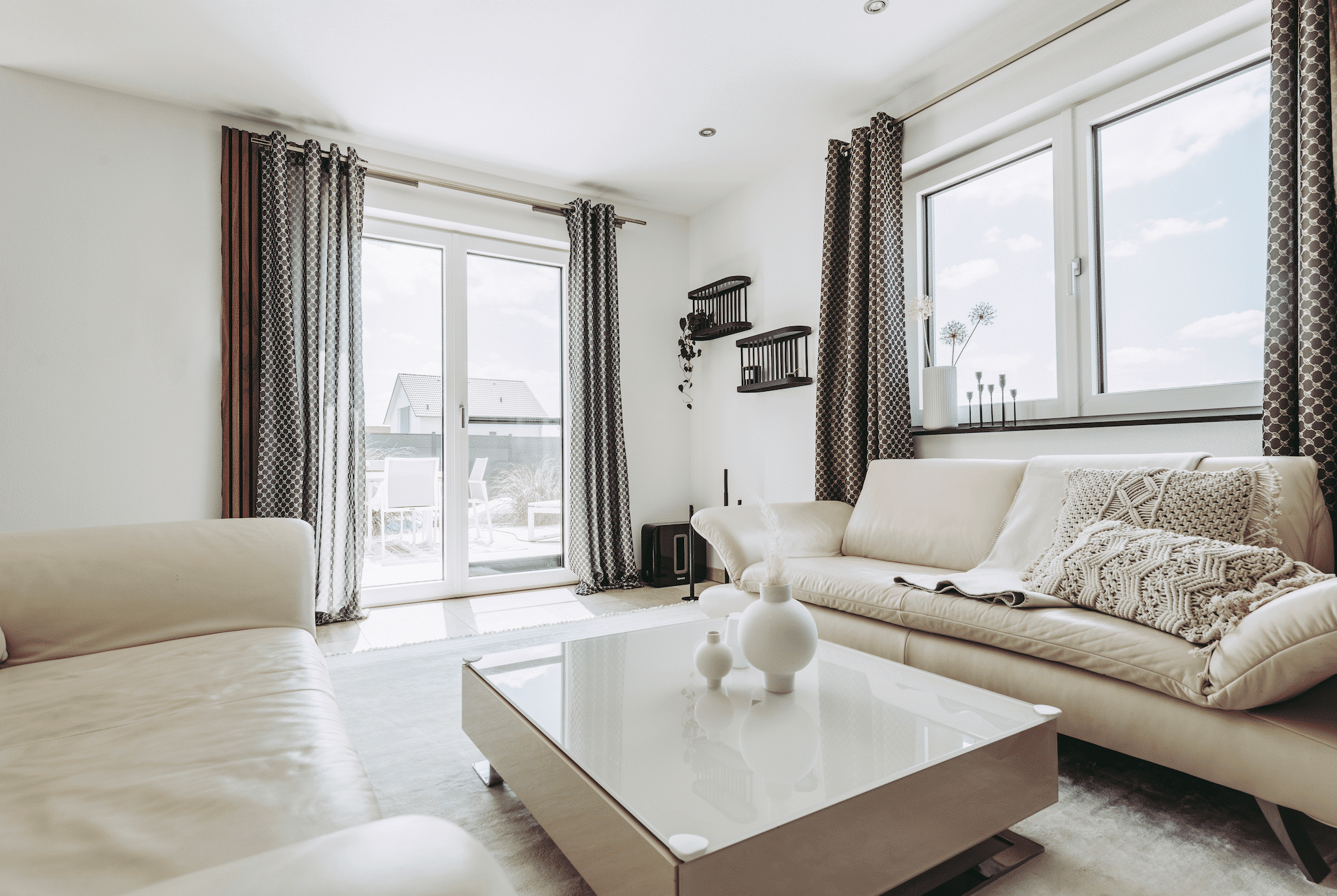 Internorm zeigt ein Wohnzimmer mit einer hellen Couch aus Leder, Kissen mit Makramee Muster, einem eckigen Beistelltisch und einer Glastüre mit Zugang zur Terrasse.