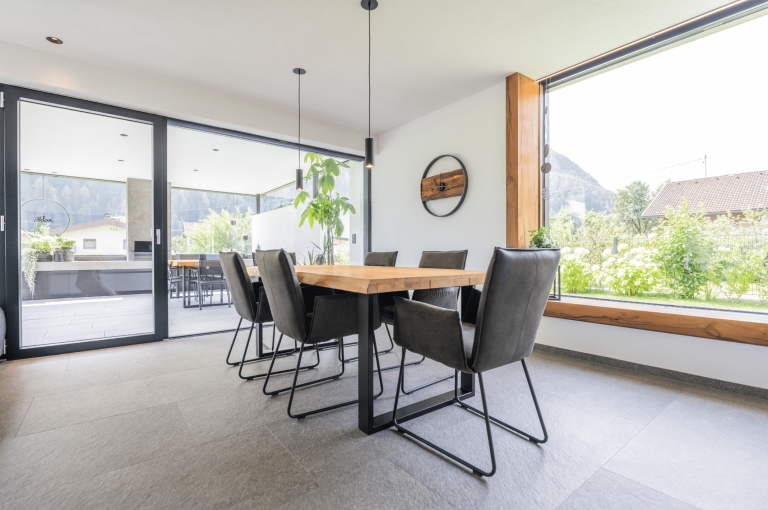 Internorm zeigt ein Esszimmer mit einem Holztisch und schwarzen Lederstühlen vor einem großen Fenster und einer Glasschiebetür mit Zugang zur Terrasse.