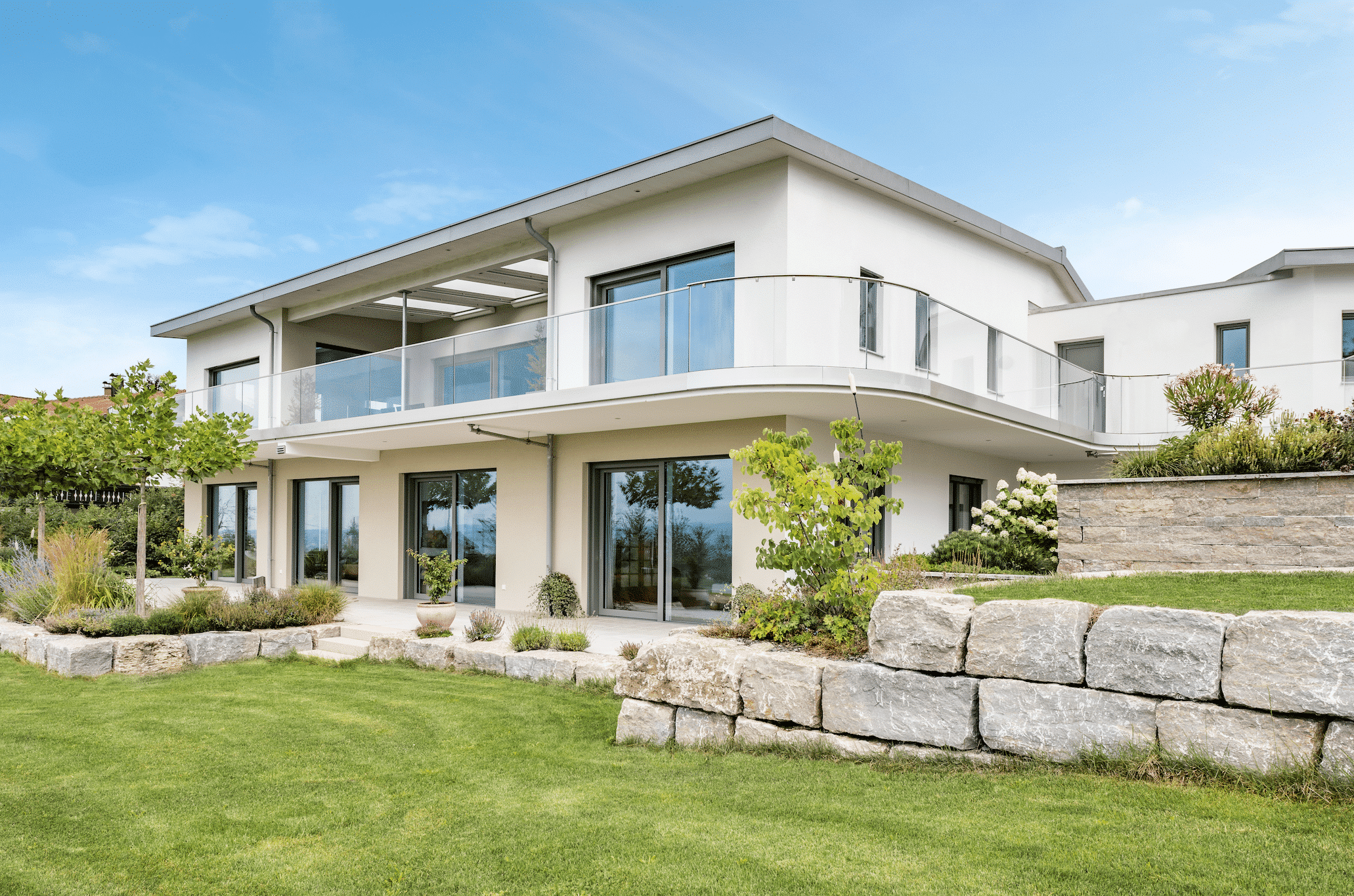 Internorm zeigt ein modernes Haus mit Flachdach und großem Balkon mit Glasgeländer und Glastüren im Erdgeschoß mit Zugang zur Terrasse und dem gepflegten Garten.