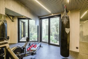 Ein moderner Fitness‑Raum mit Holzvertäfelung an den Wänden, einer Holzsprossendecke mit Leuchtröhrenund einer Terrassenschiebetür in schwarzem Alu‑Rahmen die zum Garten bzw. Atrium führt von Internorm.