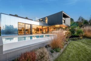 Ein moderner Villa mit Garten, Pool und einer großflächigen Wandverglasung mit schwarzem Alu-Rahmen, passend zur Fassadenverkleidung und ergänzt durch einen Dachbalkon mit Nurglasgeländer von Internorm.