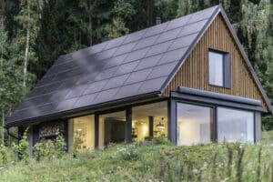 Ein kleines Haus mit Satteldach und Solardachschindeln, aussenliegendem Holzlager, und Eckverglasung und Terrassenschiebetüren schwarzem Alurahmen von Internorm.