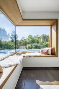 Ein ums Eck laufendes breites Sitzfenster in einer warmen Holzrahmen‑Ausführung, mit Schaffellen, Polstern und Büchern und vollem Blick auf Garten und Pool von Internorm.