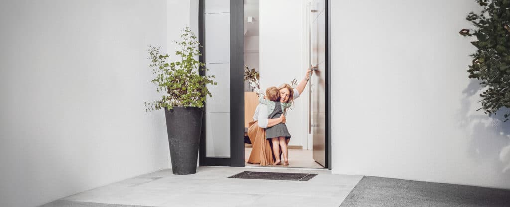 Eine Mutter empfängt ihr Kind mit einer liebevollen Umarmung an einer modernen anthrazitfarbenen Haustür mit Seitenlicht in Dreifachverglasung von Internorm.