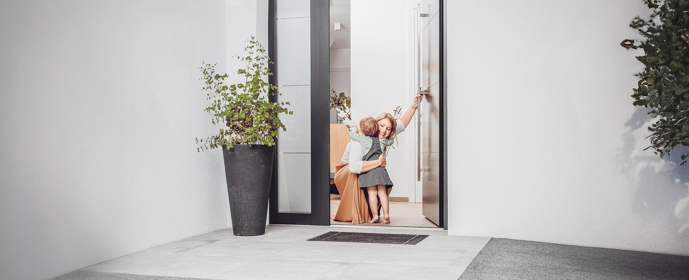 Eine Mutter empfängt ihr Kind mit einer liebevollen Umarmung an einer modernen anthrazitfarbenen Haustür mit Seitenlicht in Dreifachverglasung von Internorm.