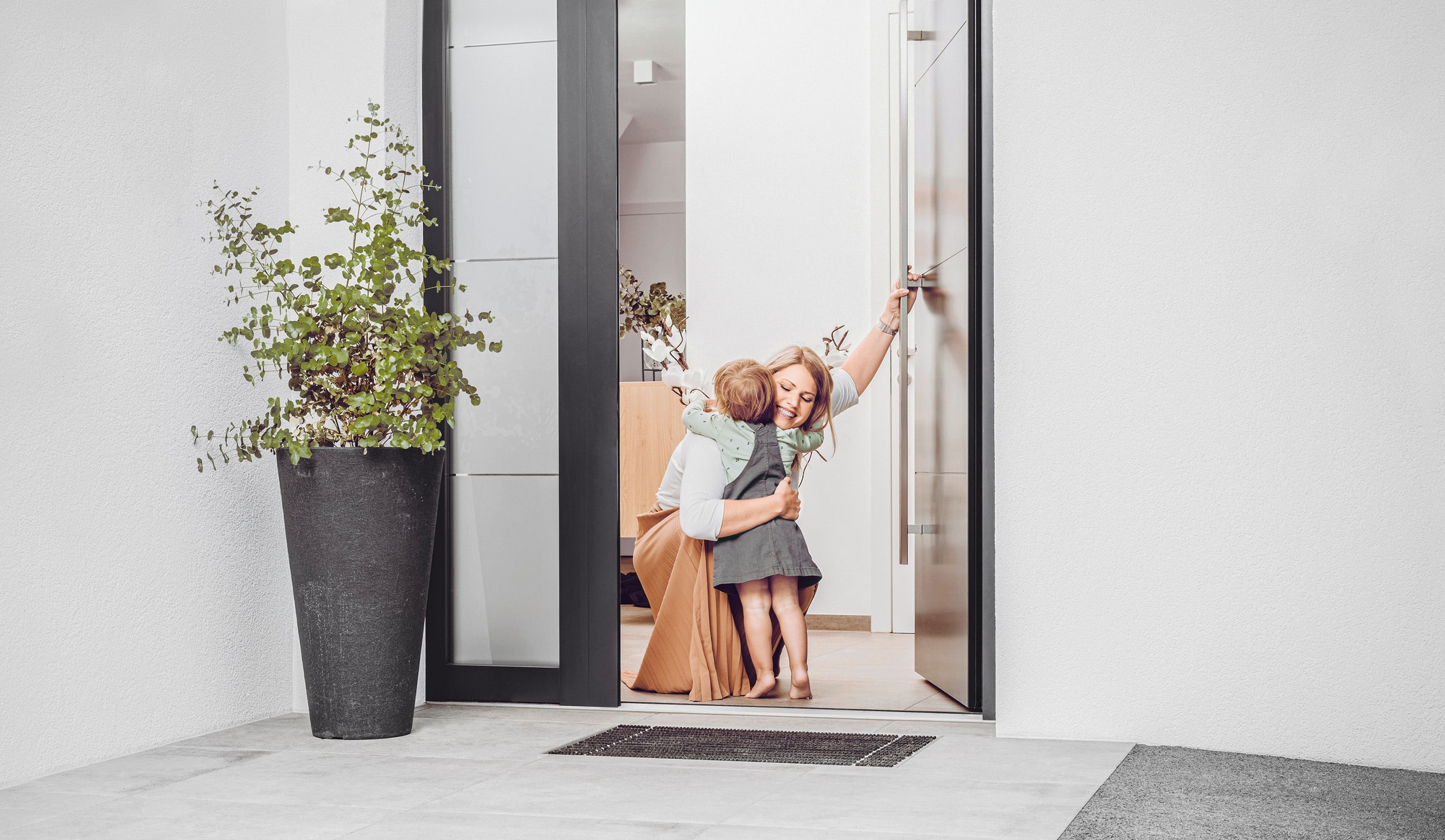 Eine Mutter empfängt ihr Kind mit einer liebevollen Umarmung an einer modernen anthrazitfarbenen Haustür mit Seitenlicht in Dreifachverglasung von Internorm.