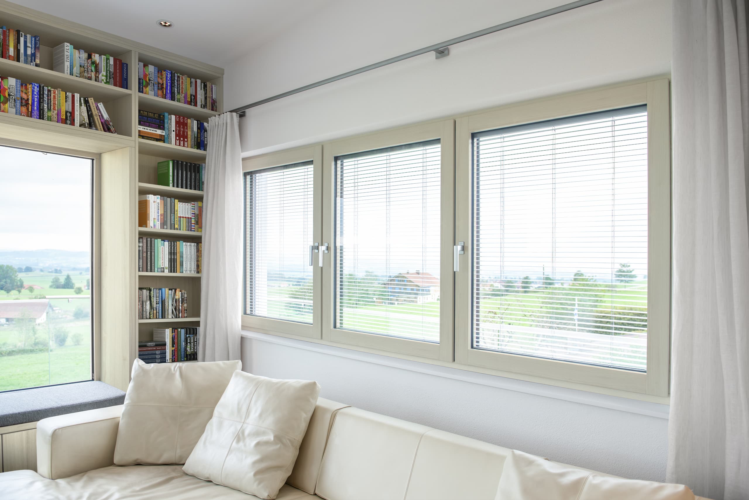 Ein heller Wohnbereich mit einem deckenhohen Bücherregalverbau mit integriertem Sitzfenster und einer 3er-Reihe sanierter Fenstern mit Außenjalousien und hellem Holzrahmen - passend zur Holzauswahl des Regals -von Internorm.