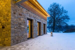 Abendliche Aussenaufnahme eines Hauses mit Steinwand, horizontalem Holzsprossen-Fassadenelement und Dachvorsprung mit LED-Schiene mit warmen Licht in winterlicher Umgebung mit Schnee und bravem Hündchen von Jahn Gewölbebau.