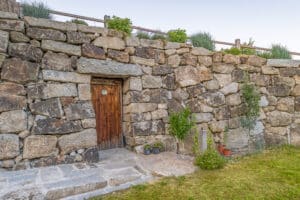 Jahn Gewölbebau zeigt eine Steinmauer an einer Böschung mit einer Türe aus Holz, die in einen Keller führt. Der Weg zum Eingang ist mit großen und kleinen Steinen verlegt.