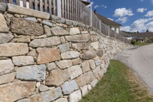 Jahn Gewölbebau zeigt eine geschwungene, schräg ansteigende Steinmauer mit Decksteinen auf der Mauerkrone und einem darauf befestigten Holzzaun.