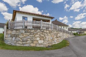Jahn Gewölbebau zeigt eine geschwungene, schräg ansteigende Steinmauer mit Decksteinen auf der Mauerkrone und einem dahinter befestigten Holzzaun vor einem Einfamilienhaus.
