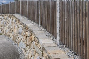 Jahn Gewölbebau zeigt eine schräg ansteigende Steinmauer mit Decksteinen auf der Mauerkrone und einem dahinter befestigten Holzzaun.