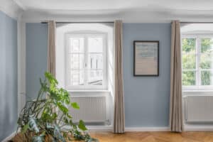 Ein Wohnraum mit weißen Kunststoff/Aluminium‑Sprossenfenstern, blauer Wandfarbe, eleganten Deckenstuck‑Elementen und Holz‑Vorhangstangen mit naturfarbenen Vorhängen von Josko.