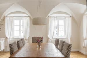 Ein Esszimmer mit Deckengewölbe und weißen Kunststoff/Aluminium‑Sprossenfenstern, seitlich drapierten weißen Vorhängen, Holzesstisch und Stühlen in einer klassischen Altbau‑Atmosphäre von Josko.