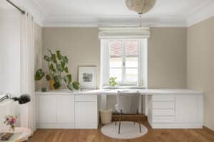 Ein Arbeitszimmer im Altbau‑Stil mit weißen Kunststoff/Aluminium‑Sprossenfenstern, innenliegenden Raffrollos, und einem eingebauten, wandabschließenden Schreibtisch vor beige Wandfarbe und klassischem Deckenstuck von Josko.