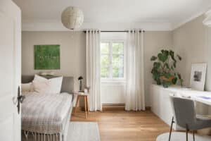 Ein Schlafzimmer im Altbau‑Stil mit weißen Kunststoff/Aluminium‑Sprossenfenstern, schwarzer Holz‑Vorhangstange, Schreibtisch und klassischem Deckenstuck von Josko.