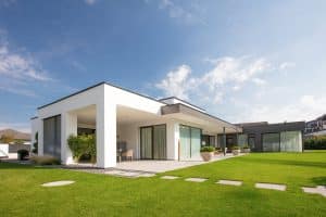 Ein moderner L-förmiger Bungalow mit Flachdach, bodentiefen Fenstern, Glaselementen und Terrasseschiebetüren, mehreren überdachten Terrassenbereichen und einem großflächigen Garten mit Trittplatten von Kickinger Bau.