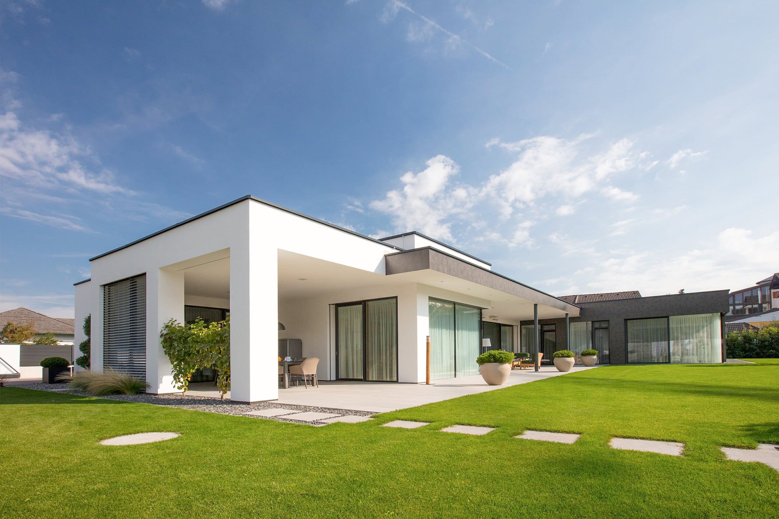 Ein moderner L-förmiger Bungalow mit Flachdach, bodentiefen Fenstern, Glaselementen und Terrasseschiebetüren, mehreren überdachten Terrassenbereichen und einem großflächigen Garten mit Trittplatten von Kickinger Bau.