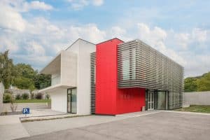 Ein modernes Firmengebäude mit Flachdach, überdachtem Balkon rotem Fassadenelement und Glasfassade mit Außenlamellen von Kickinger Bau.