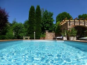 Begrünter Außenbereich eines Hauses mit einem XL-Pool, Poolumrandung aus rotem Ziegel, einer Sitzbank aus Stein und einer gemauerten Pavillon mit Holzpergola und einer weiterführenden Wand von Kickinger Bau.