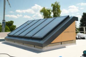 Lamilux zeigt einen schrägen Dachfenster-Aufbau mit rechteckigen Fensterflächen und dunkelgrauem Rahmen auf einem Flachdach.