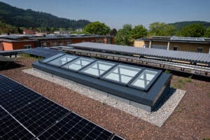 Lamilux zeigt ein schräges Dachfenster quadratischen und rechteckigen Fensterflächen und dunkelgrauem Rahmen auf einem Flachdach.