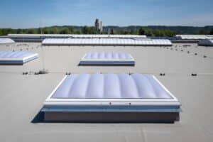 Lamilux zeigt ein langes Dachfenster mit einer Wölbung und kleinere Dachfenster mit welligem Milchglas auf einem Flachdach.